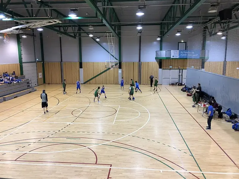 Interiör i en kommunal sporthall med parkettgolv, pågående match och åskådare längs väggen